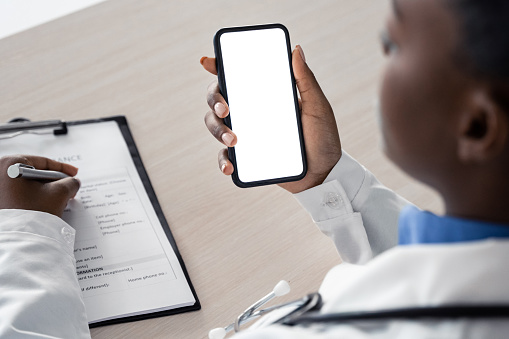 African american female doctor holding using cell phone app mock up white screen, over shoulder closeup view. Healthcare telemedicine online remote consultation mobile medical tech application concept