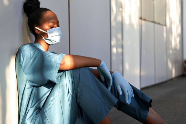 Tired exhausted female african scrub nurse wears face mask blue uniform gloves sits on hospital floor. Depressed sad black ethic doctor feels fatigue burnout stress, lack of sleep, napping at work. Tired exhausted female african scrub nurse wears face mask blue uniform gloves sits on hospital floor. Depressed sad black ethic doctor feels fatigue burnout stress, lack of sleep, napping at work. exhaustion stock pictures, royalty-free photos & images