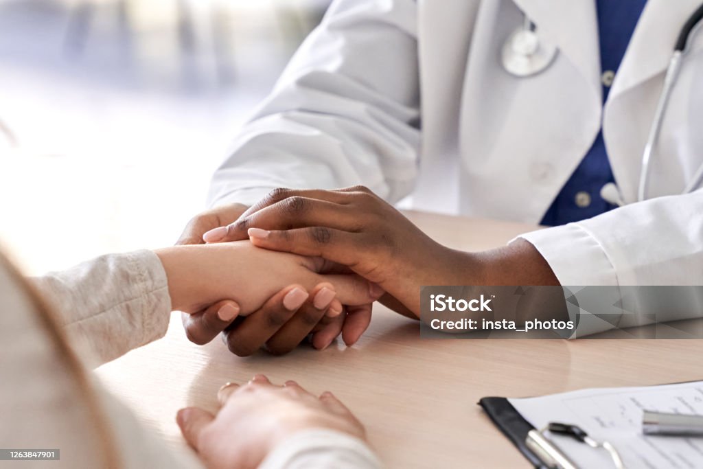 African female doctor hold hand of caucasian woman patient give comfort, express health care sympathy, medical help trust support encourage reassure infertile patient at medical visit, closeup view. Doctor Stock Photo