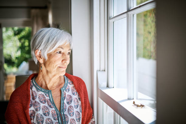istock Senior woman standing alone by a window at home 1263853625