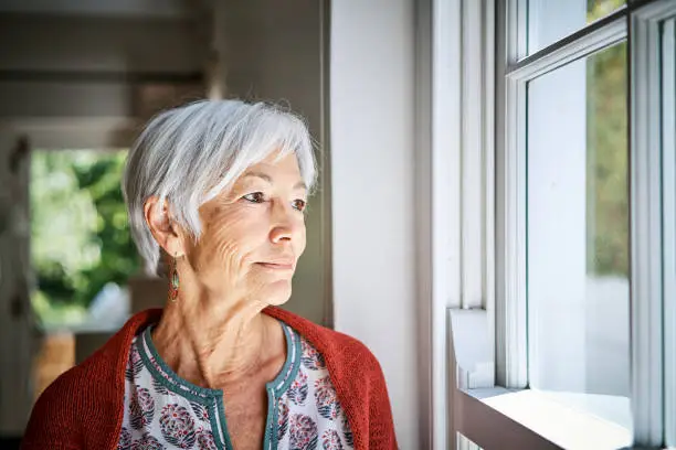 Senior woman standing alone in her living room at home and looking out through a window