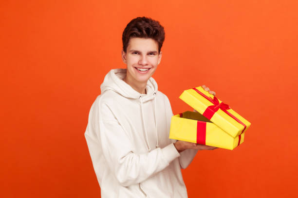 adolescente sorridente satisfeito em moletom casual com capuz desfrutando de segurando caixa de presente e olhando para a câmera, melhor loja presente, anúncio - hood men portrait profile - fotografias e filmes do acervo