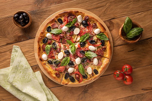 Colorful Italian pizza with ham, salami, black olives, mozzarella cheese, red tomatoes and basil leaves on natural wooden background and few ingredients in wooden bowls. Linen napkin on side. Flat lay