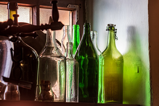 Antique multi-colored glass bottles sit on the top shelf near the dusty window. Wine bottles of various shapes and colors. Whimsical shadows.