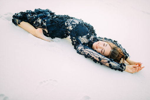 Woman in lace black dress lies in desert on white sand, relaxes and enjoys.