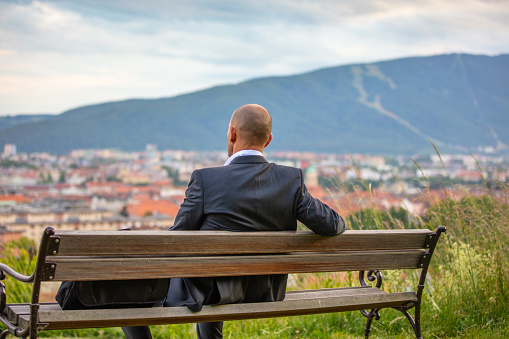 Businessman sitting on bench with his computer bag and looking at city from hill
