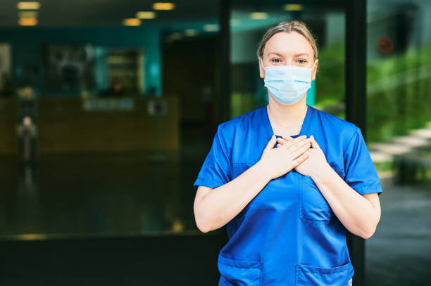 Young female scrub nurse wear blue uniform and face mask, standing in hospital entrance stock photo