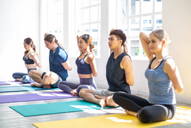ダイバーシティアジアと白人の若者のバナーグループは、フィットネススタジオでクラスでヨガカウフェイスポーズ(gomukhasana)を練習するマットの上に座っています。健康的な健康ライフスタ - yoga class caucasian young adult group of people ストックフォトと画像