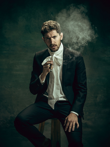 Smoking cigar. Young man in suit as Dorian Gray isolated on dark green background. Retro style, comparison of eras concept. Beautiful male model like classic literature character, old-fashioned.