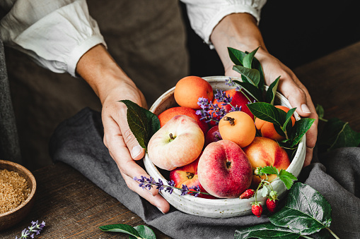 Hand of woman holding a bowl of fresh fruits. Ripe peaches, Apricots, strawberries and apples with leaves in ceramic bowl over wooden table.
