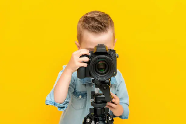 Adorable little kid taking a photo using a digital camera on a tripod isolated on a yellow background