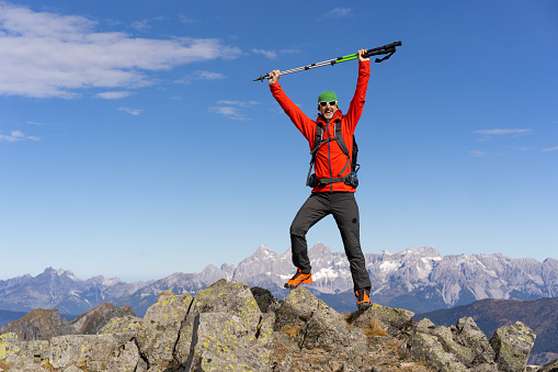 successful happy smiling mature man hiker mountaineer standing on mountain peak with hiking poles raised up, beautiful panorama of alpine mountains in background