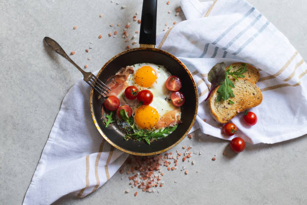 istock Pan of fried scrambled eggs, bacon, greens and cherry-tomatoes with bread on grey stone table, top view 1263906219