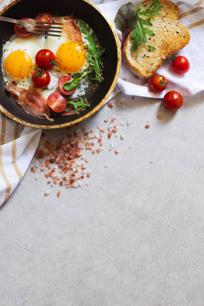 istock Pan of fried scrambled eggs, bacon, greens and cherry-tomatoes with bread on grey stone table, top view 1263906256