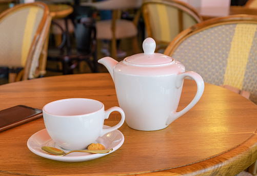White porcelain teapot, tea cup and smartphone stand on a table in a cafe. Horizontal orientation, selective focus.