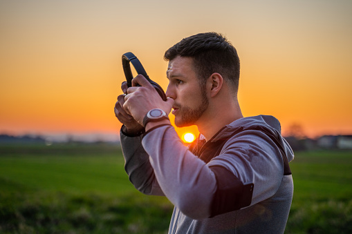 Side view of man in sportswear putting on headphones in rural surrounding at sunset