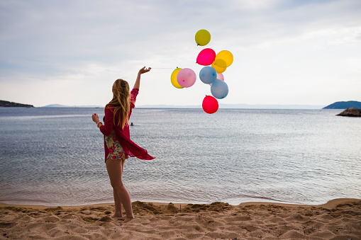 Casually dressed young woman with colorful balloons walking at the beach on a lovely sunny day during her summer vacation in Greece