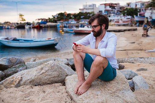 Young bearded man sitting at the beach, relaxing and using headphones and a smartphone on a lovely day in Greece