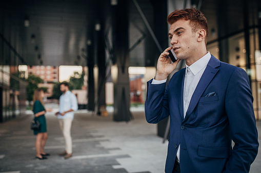 Young businessman standing in front of his office building and using smart phone. Two young business people talking in background.