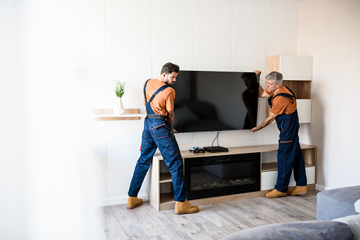 Two handymen, workers in uniform hanging, installing tv television on the wall indoors. Repair and assembly service concept. Selective focus. Horizontal shot