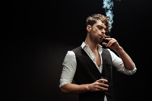 Man in shirt and waistcoat smoking cigar and holding glass of whiskey isolated on black