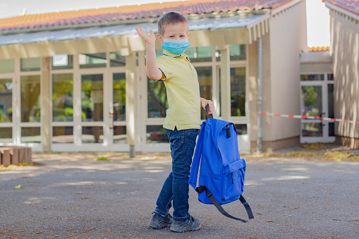 A boy with a blue backpack and wearing a mask waves goodbye in front of the school or kindergarten door. Children are happy to return to school after the epidemic. The new school year. Back to school.