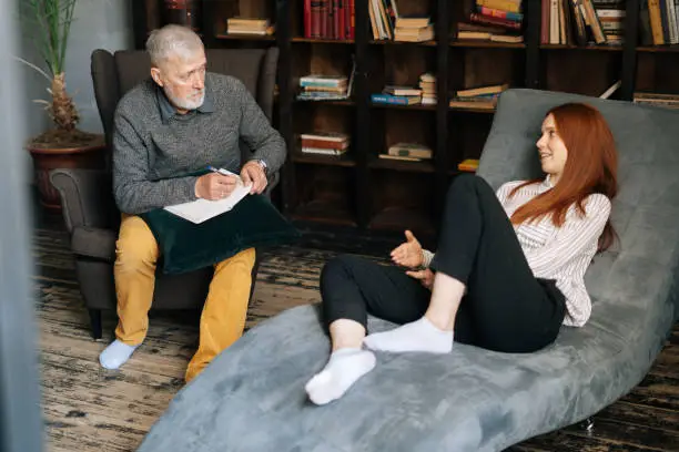 Bearded mature man psychologist with gray-hair is supporting and consoling depressed patient. Professional senior psychotherapist is giving healthcare advice to patient and taking notes.