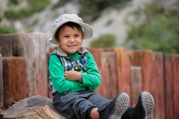 Little boy with arms crossed and sitting, waiting at the trailhead wearing his hiking boots and hat. stock photo