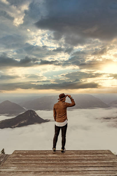 Young man standing on top of cliff in summer mountains at sunset and enjoying view of nature