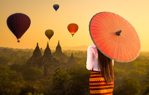 A young Burmese woman is walking with a red umbrella. Young girl and Sunrise many hot air balloon with stupas in Bagan, Myanmar. space for text.