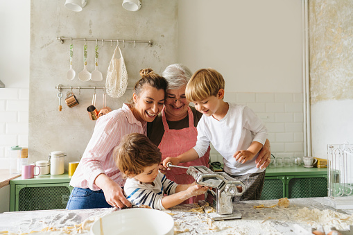 Photo of cute little boys learning how to make homemade pasta, assisted by their grandmother and mother in the kitchen; rolling out the pasta dough using the manual pasta maker; having a great time while making food and enjoying each other's company.