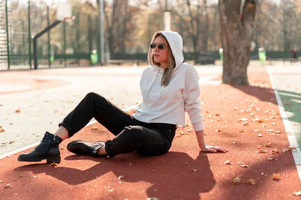 Outdoor portrait of young beautiful woman with long in sunglasses and a white hooded sweater sitting on the sportsground track.  youth culture summer pastime
