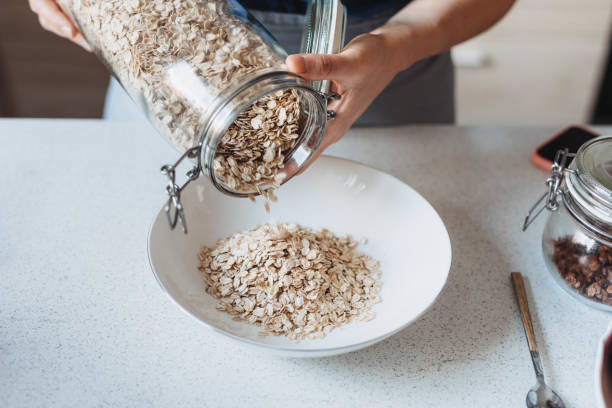 fare una colazione sana e nutriente: mani di una donna che versa avena arrotolata in una ciotola - oat oatmeal rolled oats oat flake foto e immagini stock