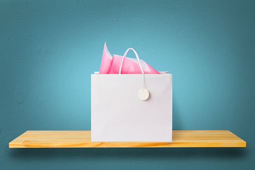 Blank white shopping bag with pink paper decoration on a wooden shelf against blue wall.