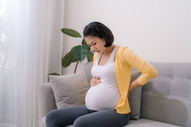 азиатская беременная женщина имеет боль в спине и чувствовать себя подавленным, сидя на диване - backache pain women illness стоковые фото и изображения