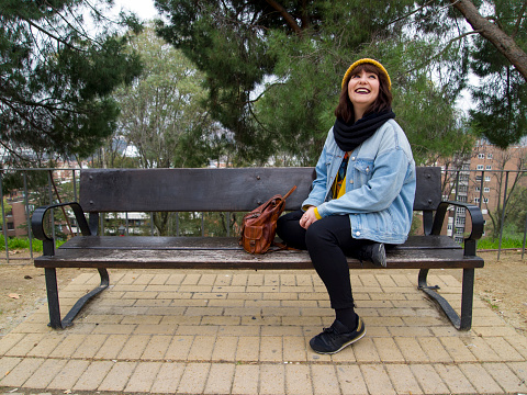 Young woman with yellow woolen cap sitting on a bench looking up and smiling. Autumn urban concept.