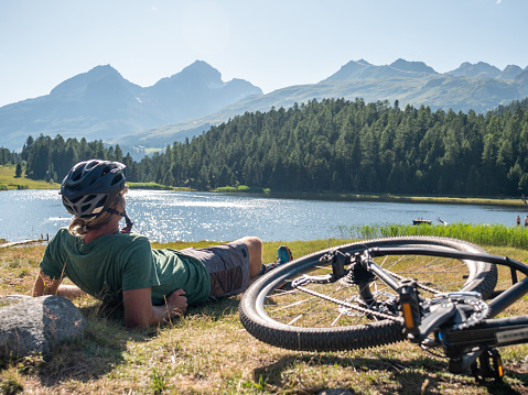 Mountain biker resting on grass area near alpine lake enjoying break in the sun. Man with bicycle enjoying nature and mountain range landscape in summer, outdoor activities