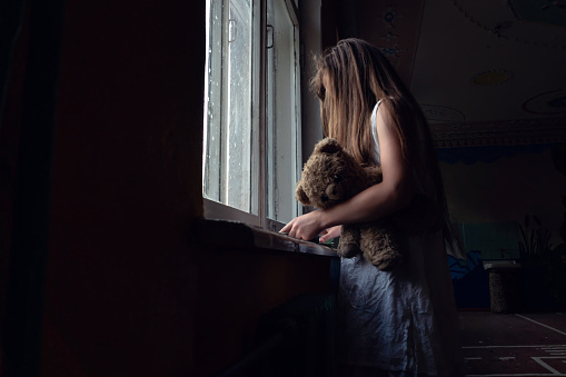 girl in a white dress with long hair and a Teddy bear under her arm stands at the window and reads a book in the dark. Concept of horror, mysticism