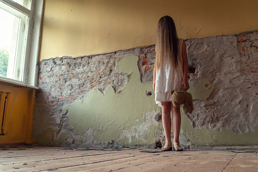 girl in a white dress with long hair and a Teddy bear in her hands stands near a ruined wall inside an abandoned building. Concept of horror, mysticism