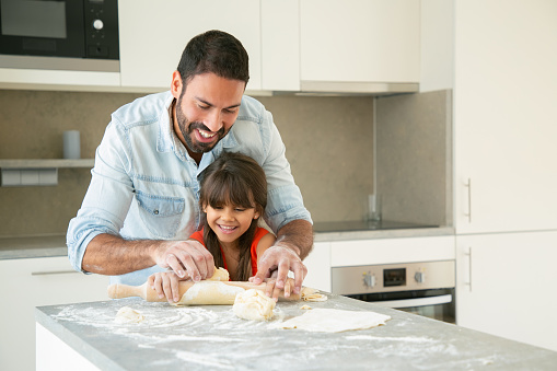 Joyful happy dad and his girl enjoying time together while rolling and kneading dough in kitchen. Father helping daughter to bake bread or pies. Home activities concept