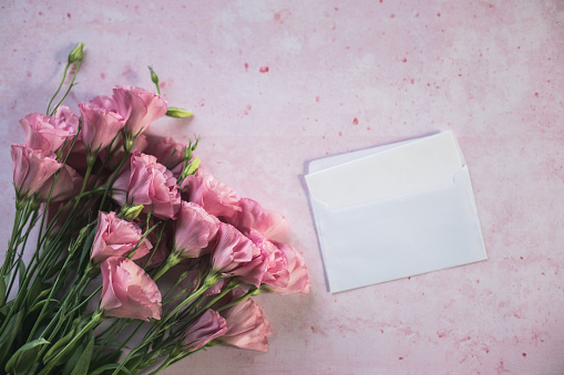 Directly above shot of envelope with letter in it next to a beautiful bouquet of pink lisianthus flowers against a pastel pink background.