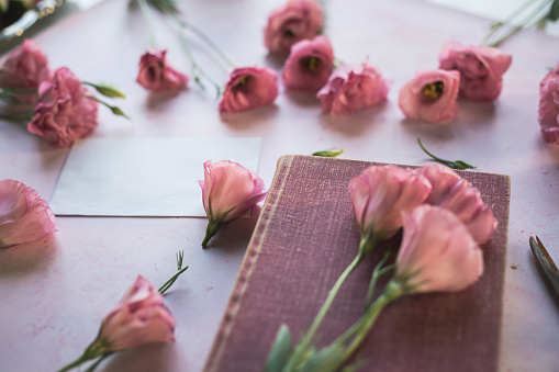 Selective focus of a pink book decorated with pink lisianthus flowers and also scattered around an envelope. All that is arranged on a pastel pink background.