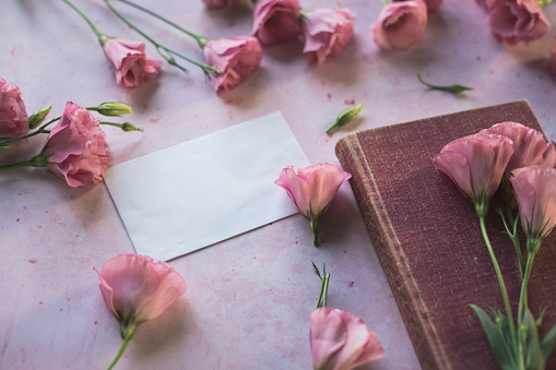 Flat lay of abundance of scattered pink lisianthus flowers around an envelope and a book next to it.