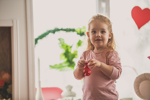 Front view, copyspace of cute little girl standing in the living room and holding a red balloon before blowing it up.
