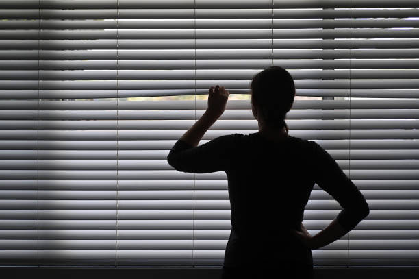 Suspicious woman looking outside through a venetian curtain blinds Suspicious adult woman looking outside home window from behind Venetian curtains. Concept photo of person that have Pistanthrophobia and Agoraphobia, a person who don't trust fear of leaving the house. dark window blinds stock pictures, royalty-free photos & images