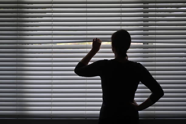 Suspicious woman looking outside through a venetian curtain blinds Suspicious adult woman looking outside home window from behind Venetian curtains. Concept photo of person that have Pistanthrophobia and Agoraphobia, a person who don't trust fear of leaving the house. peeking stock pictures, royalty-free photos & images