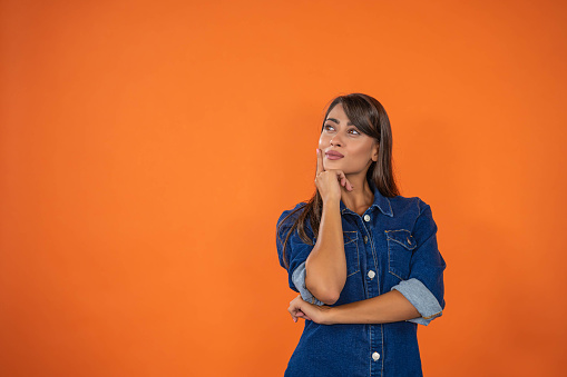 Thoughtful  brunette with denim dress looking at blank text space, orange background. Concept of shopping, summer sale. Online shopping concept, copy space provided. The scene is situated in controlled studio environment in front of orange background. Photo is taken with SONY A7III camera.