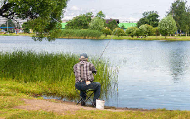 Elderly man sits on a folding chair on the shore of a city pond and fishes stock photo