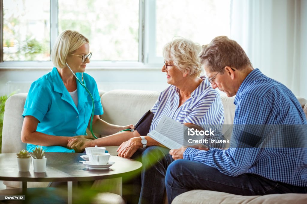 Home nurse checking the blood pressure of the elderly lady Female home nurse measuring the arterial pressure of the senior woman. An elderly lady and her husband sitting in a living room. Parkinson's Disease Stock Photo