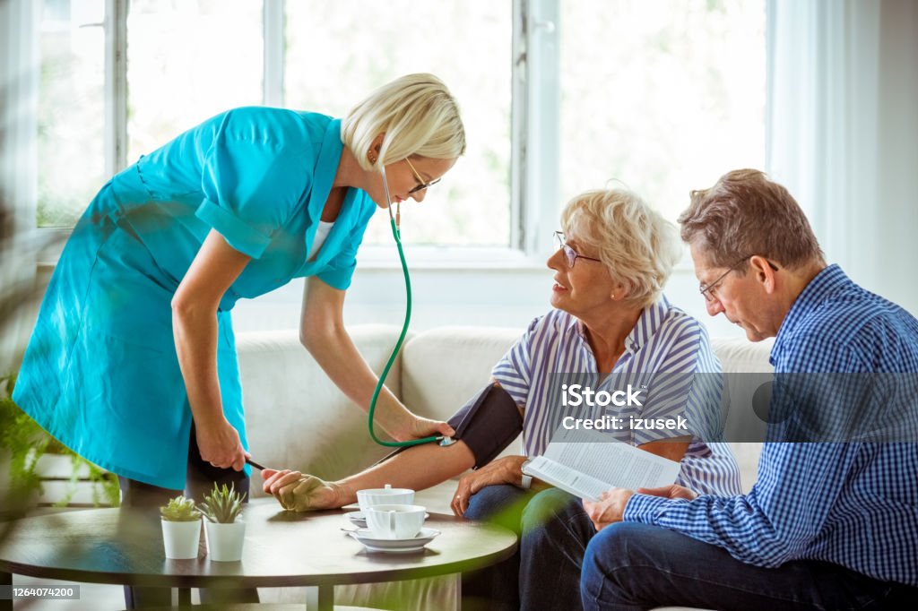 Home nurse checking the blood pressure of the elderly lady Female home nurse measuring the arterial pressure of the senior woman. An elderly lady and her husband sitting in a living room. Blood Pressure Gauge Stock Photo